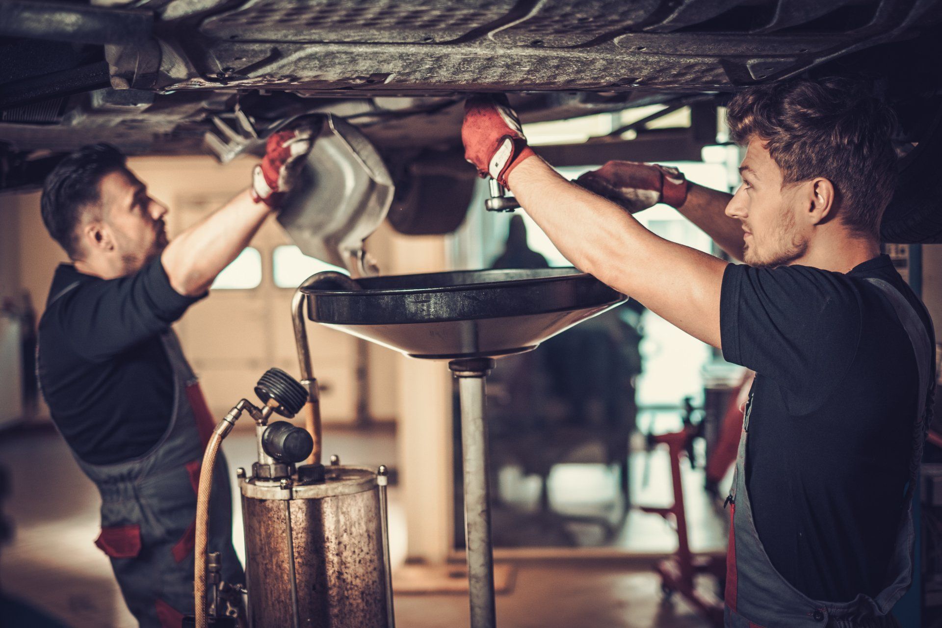 Two mechanics in a garage working together to drain fluid from the underside of a car into a collection funnel.