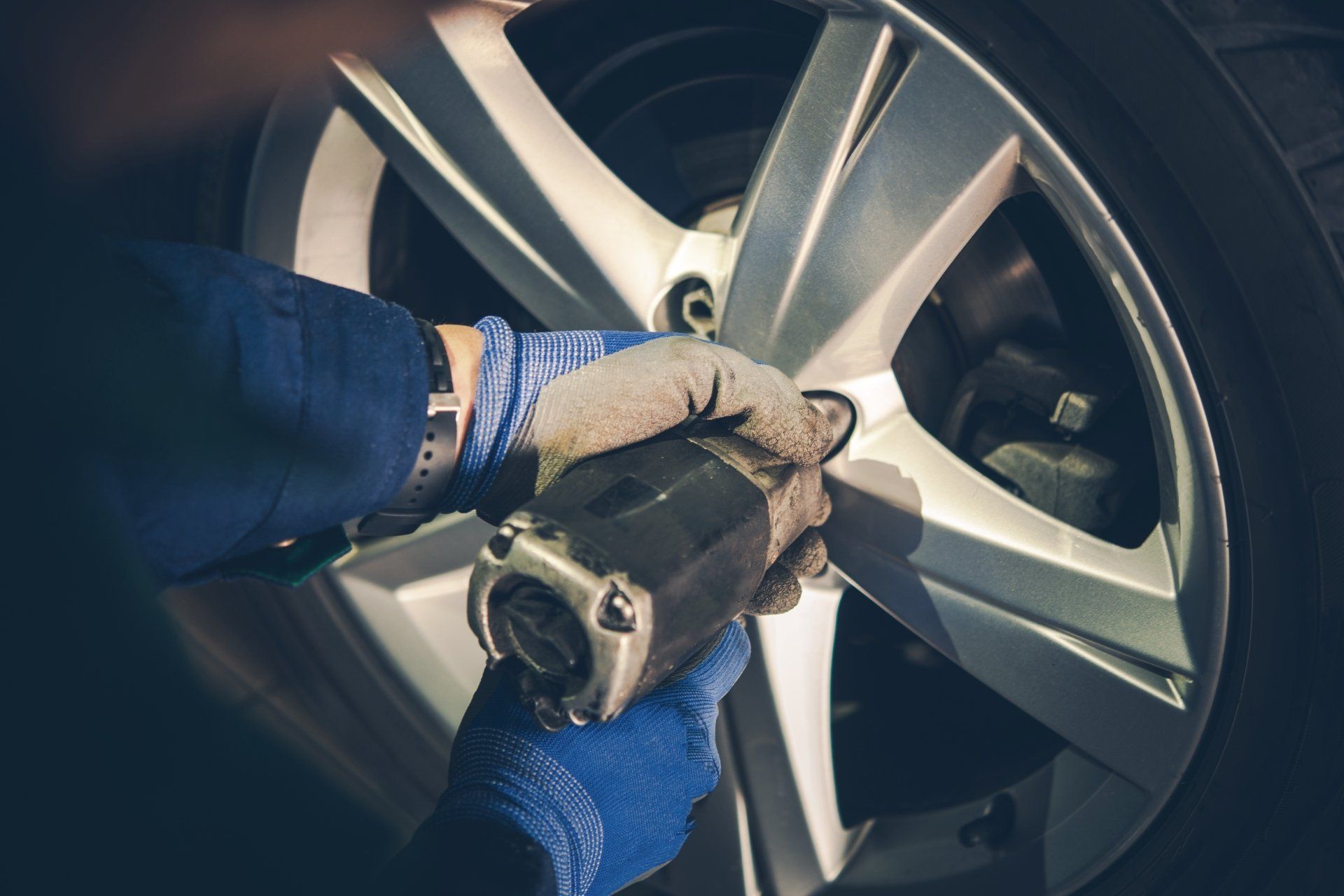 A mechanic wearing blue gloves uses a power tool to tighten lug nuts on a car's silver alloy wheel.