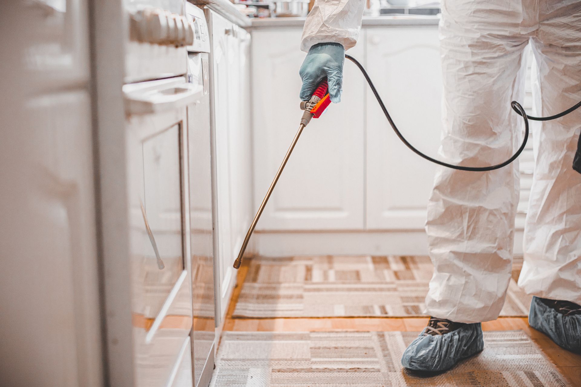 Pest control services technician spraying under kitchen cabinets in protective suit