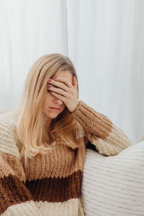 Woman seated on a couch, covering her face with one hand in a softly lit room.