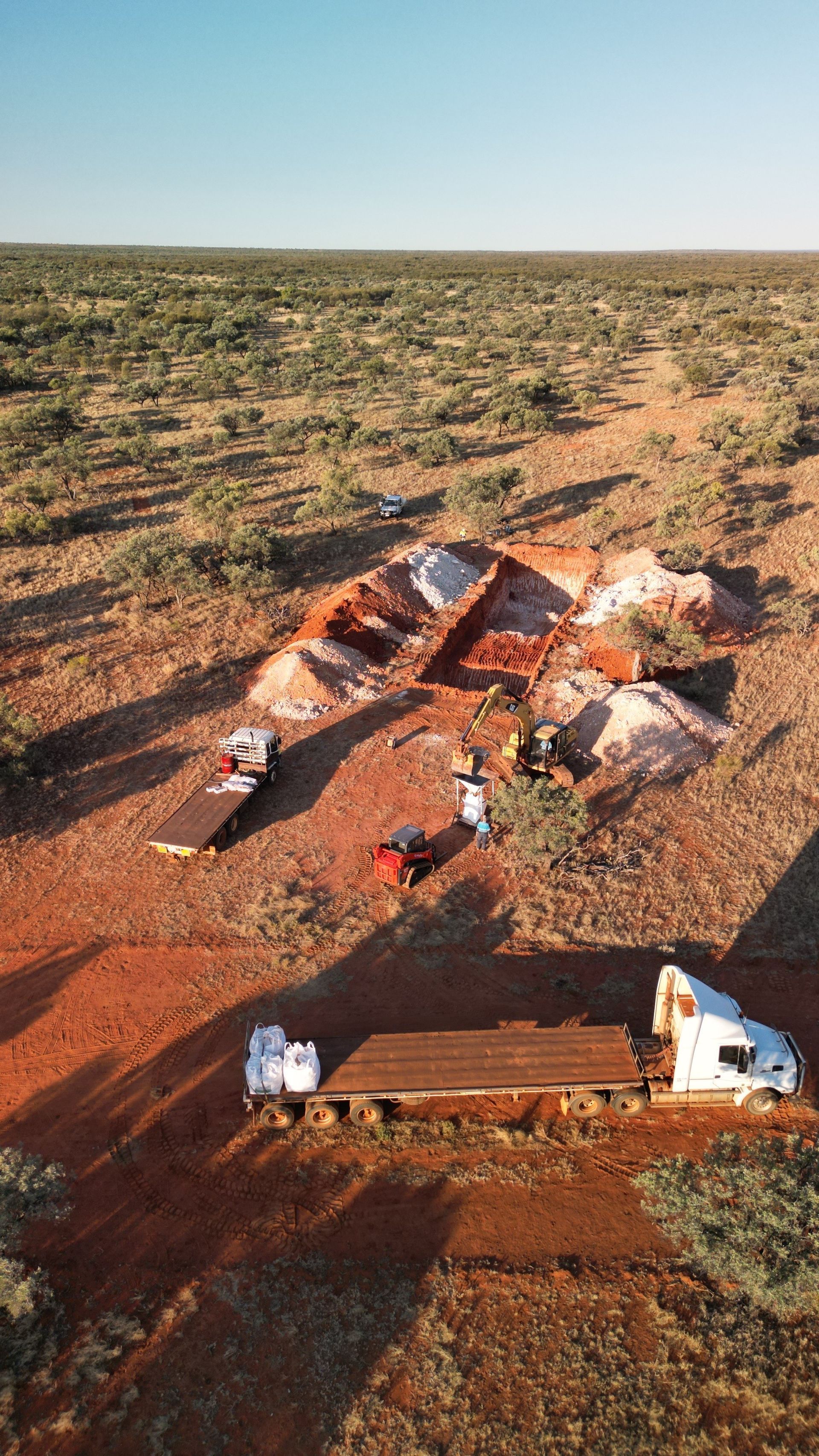 Trucks near piles of reddish soil in an arid, open landscape under a clear blue sky.