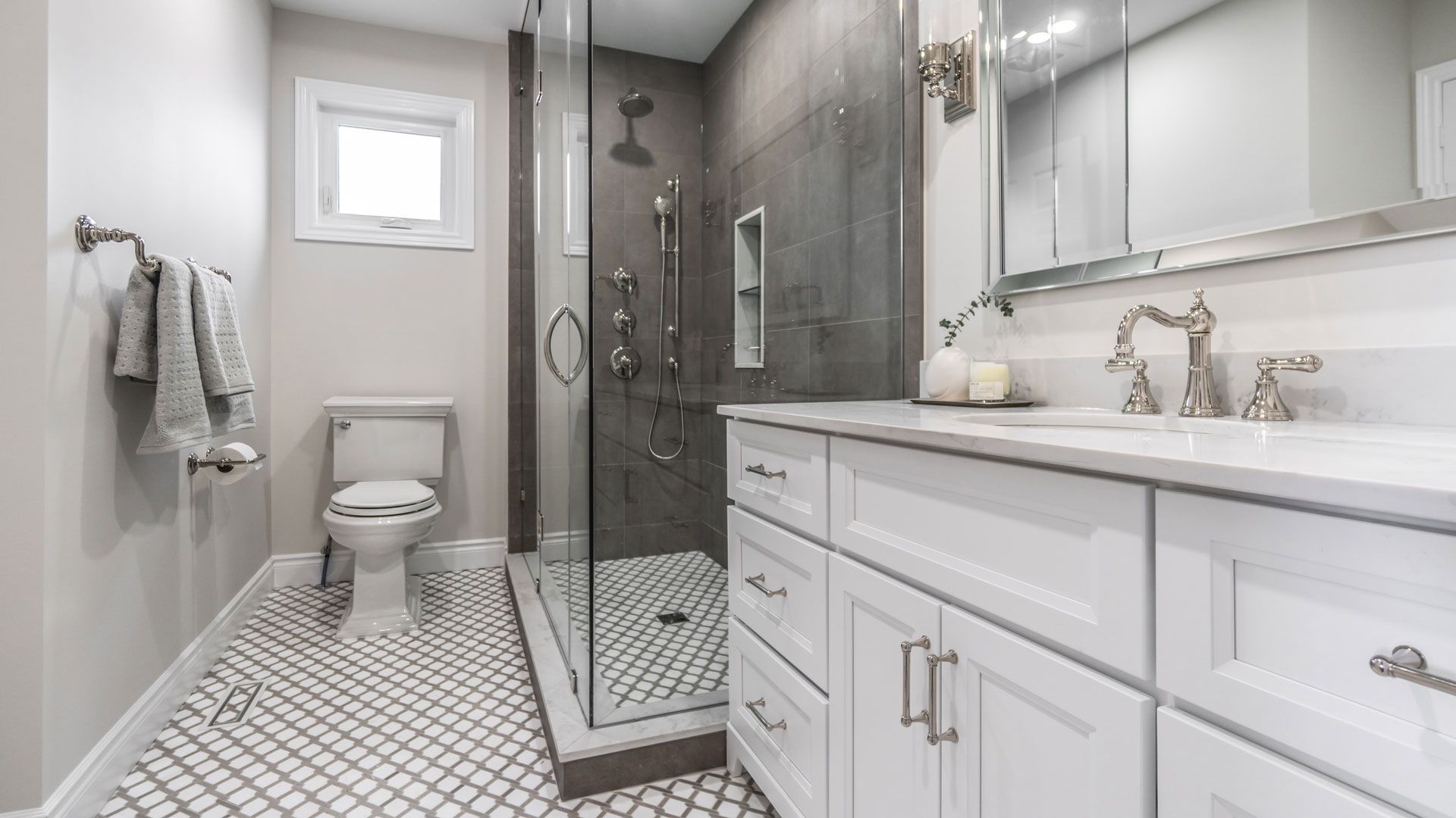 White bathroom with a glass shower, toilet, and vanity with a grey tiled shower wall.