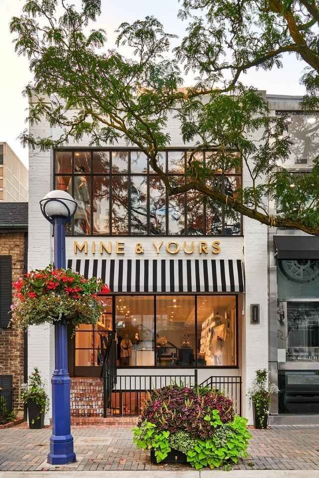 Storefront of “MINE & YOURS” with large windows, hanging flowers, and a blue lamppost on a tree-lined street