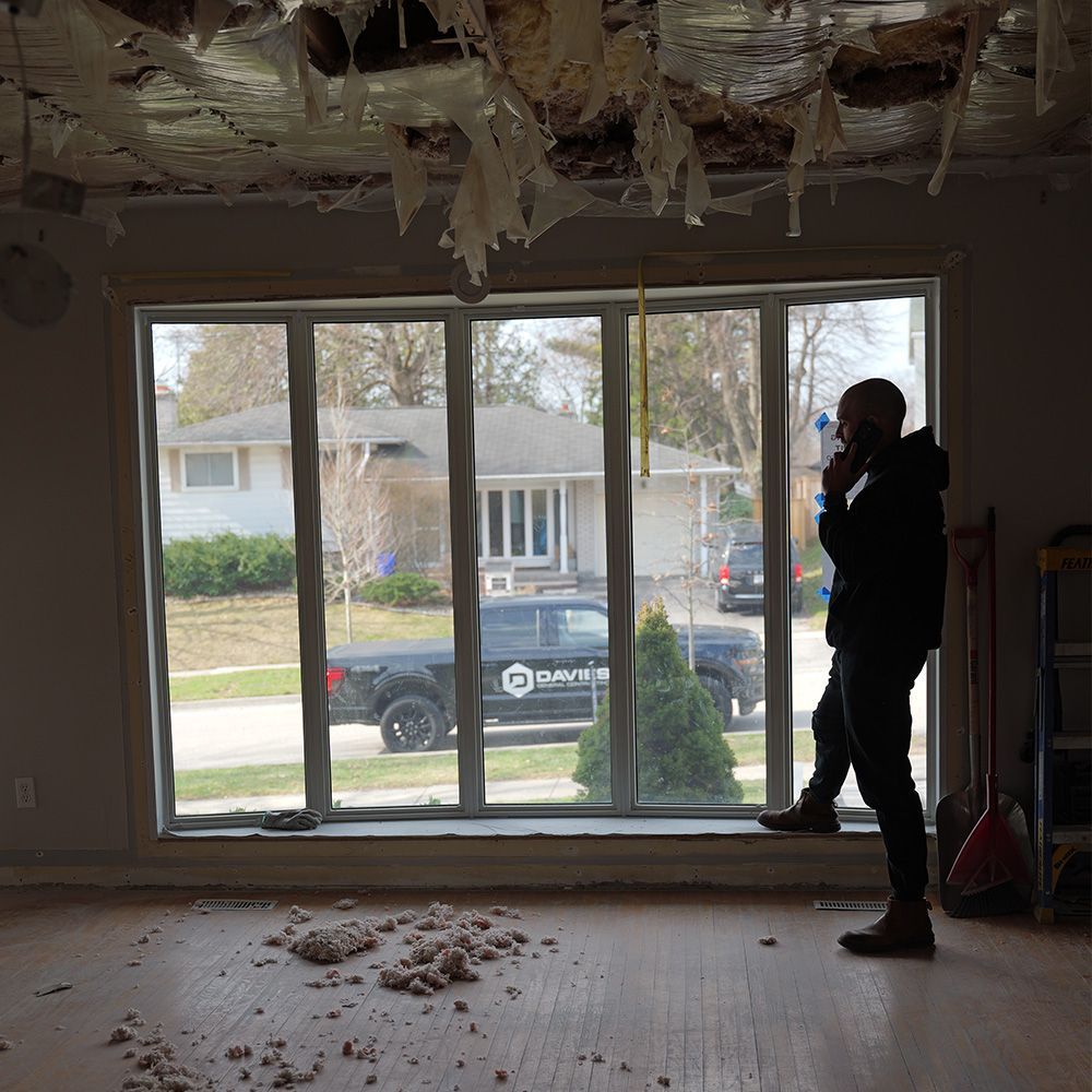Person in a damaged room with exposed ceiling insulation, looking out a large window at a suburban street.