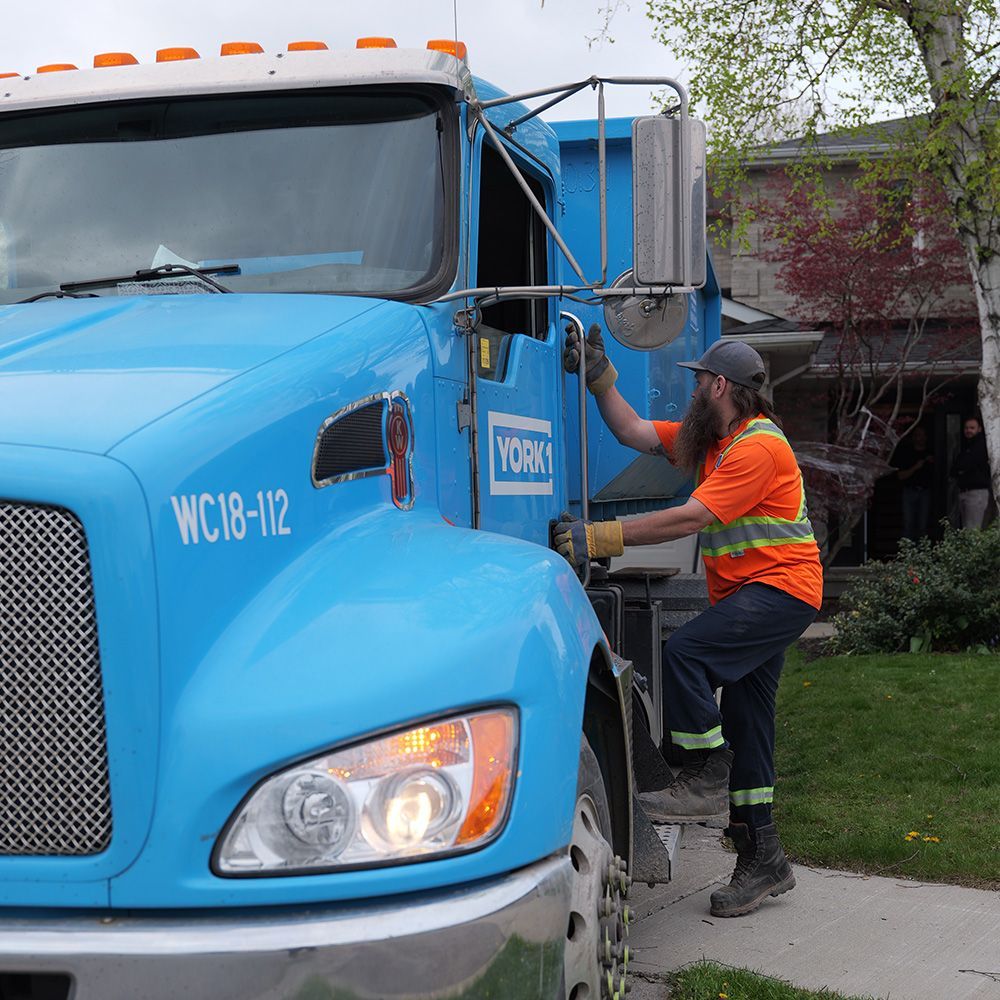 Worker in orange vest climbing into a blue garbage truck beside a house