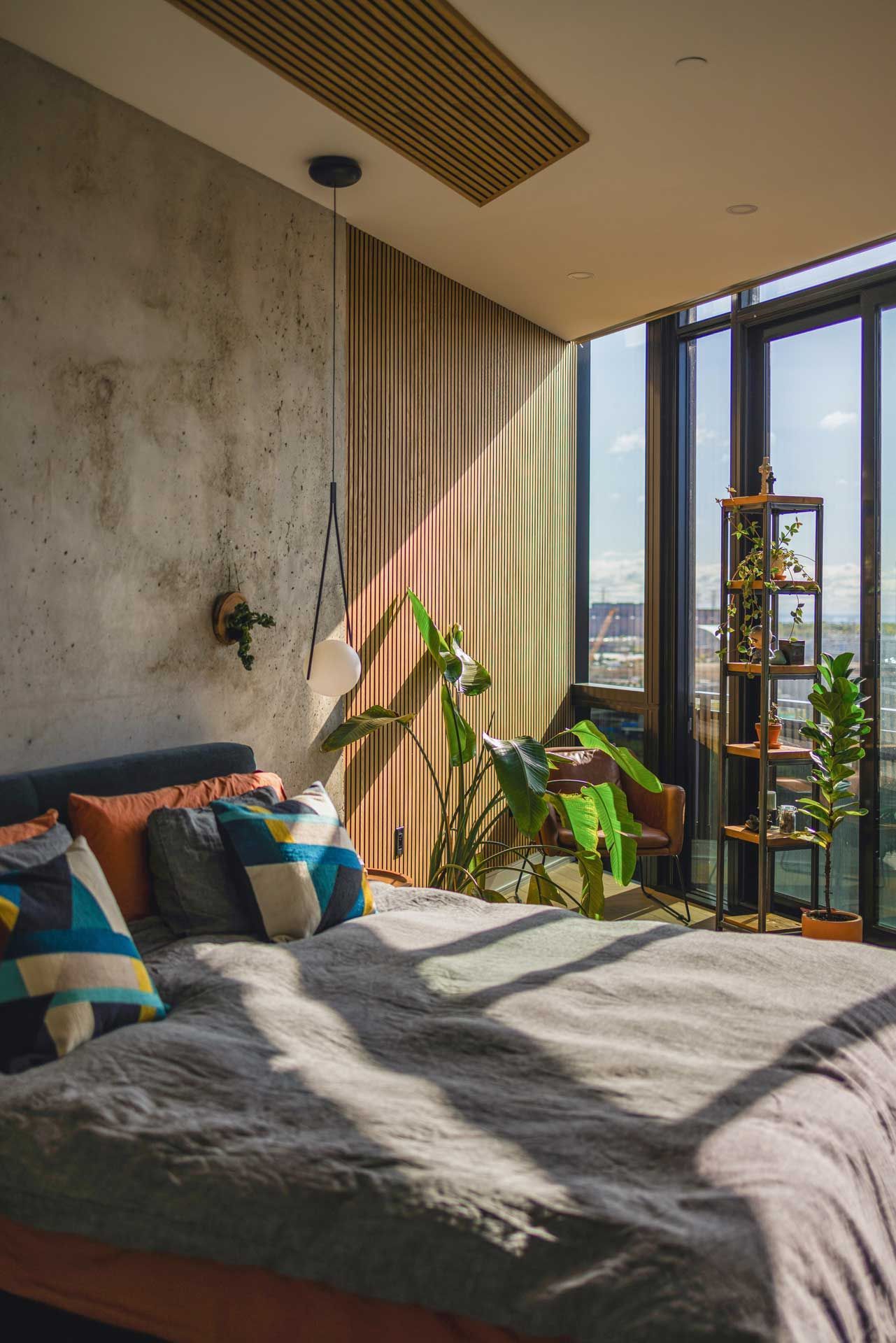 Bedroom with bed, plants, sunlight streaming through window, and exposed concrete wall.