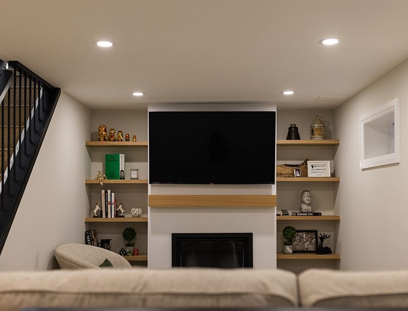 Living room with fireplace, TV, and built-in shelves. Light walls, black stairs.
