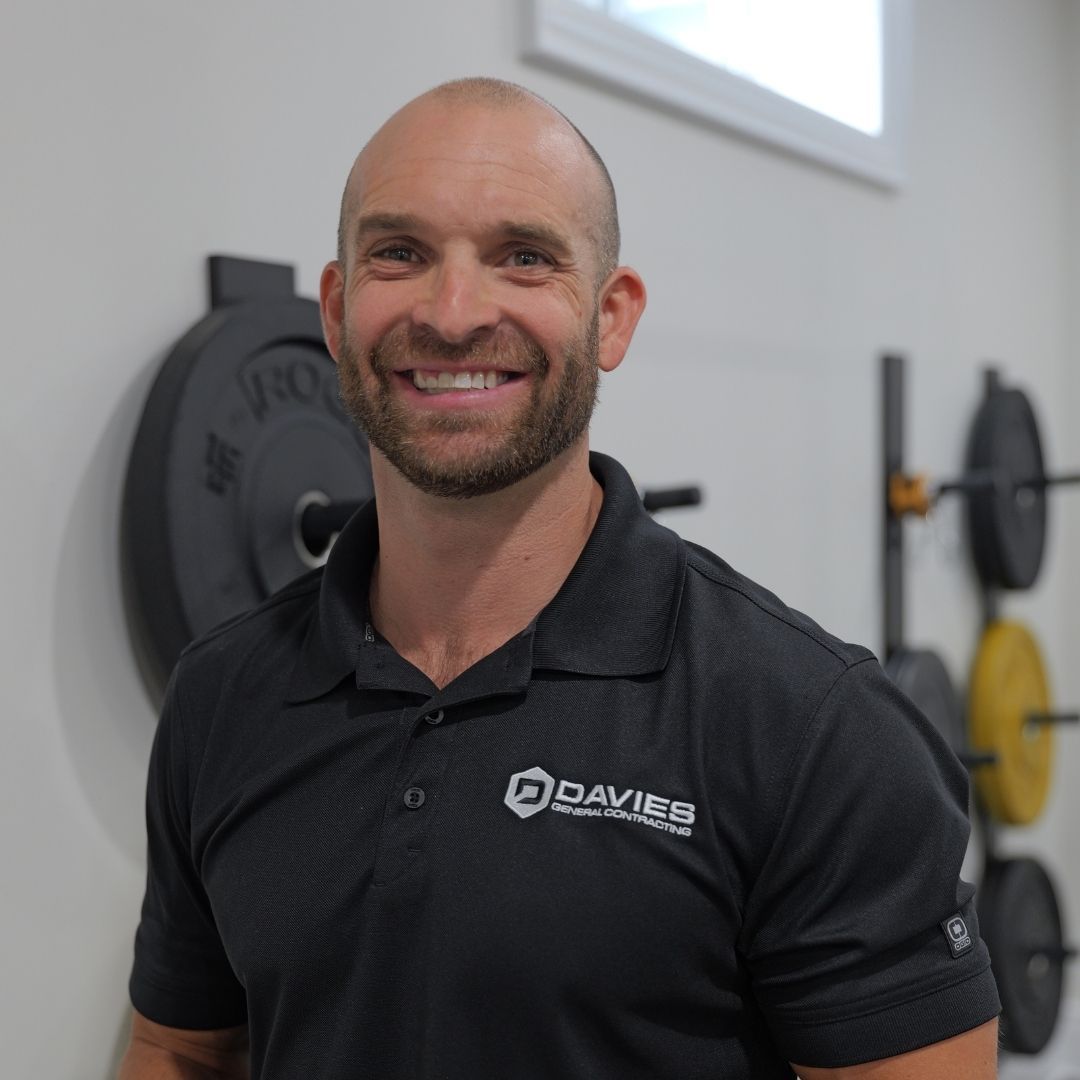 Smiling man in a black polo standing in a gym with weight plates and equipment behind him
