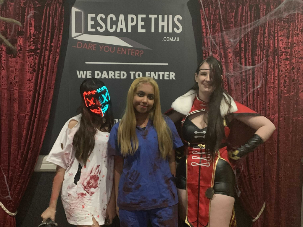 Three people in halloween costumes are posing for a picture in front of a sign that says escape this