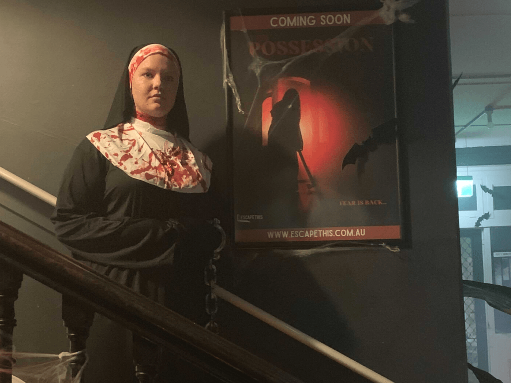 A GM dressed as a bloodstained nun is standing in front of a poster for The Possession Escape Room.