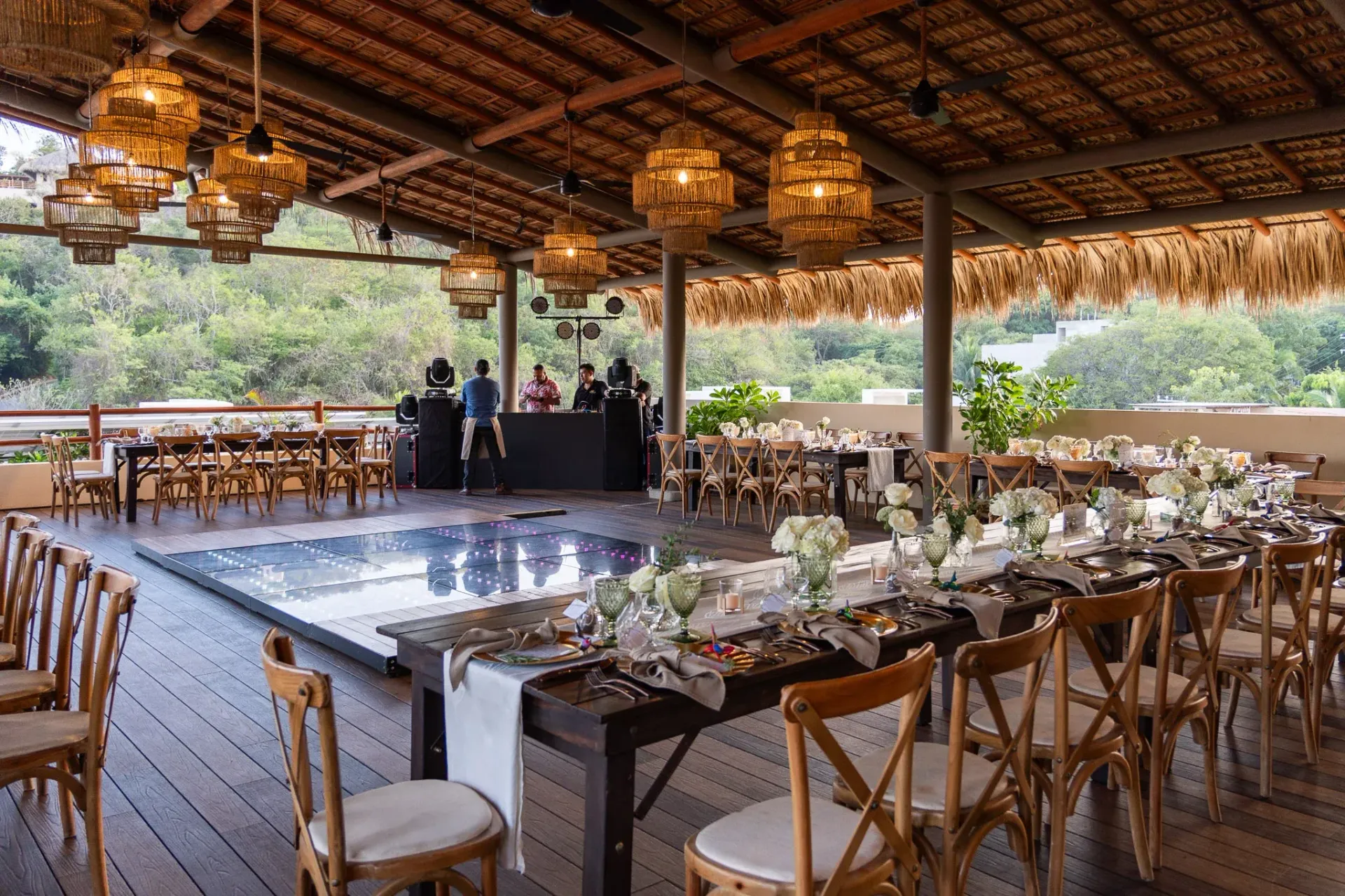 Espacio para eventos al aire libre con mesas, sillas, pista de baile y escenario bajo techo de paja.