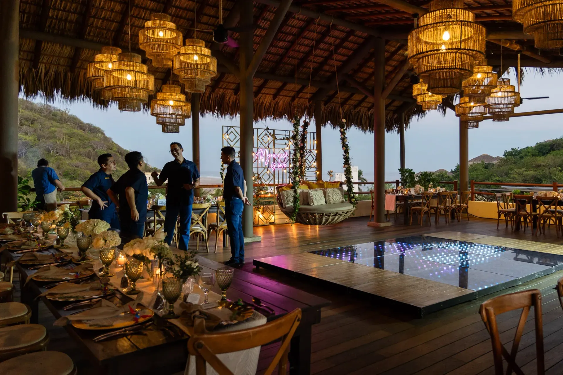 Montaje de un salón de recepción de boda en un pabellón de estilo tropical con mesas, pista de baile y luces colgantes.