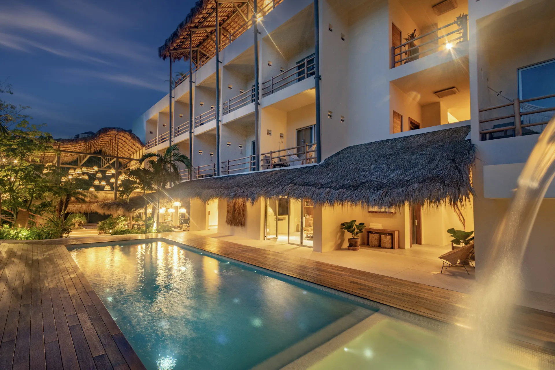 Hotel de noche con piscina, habitaciones iluminadas y techo de paja.