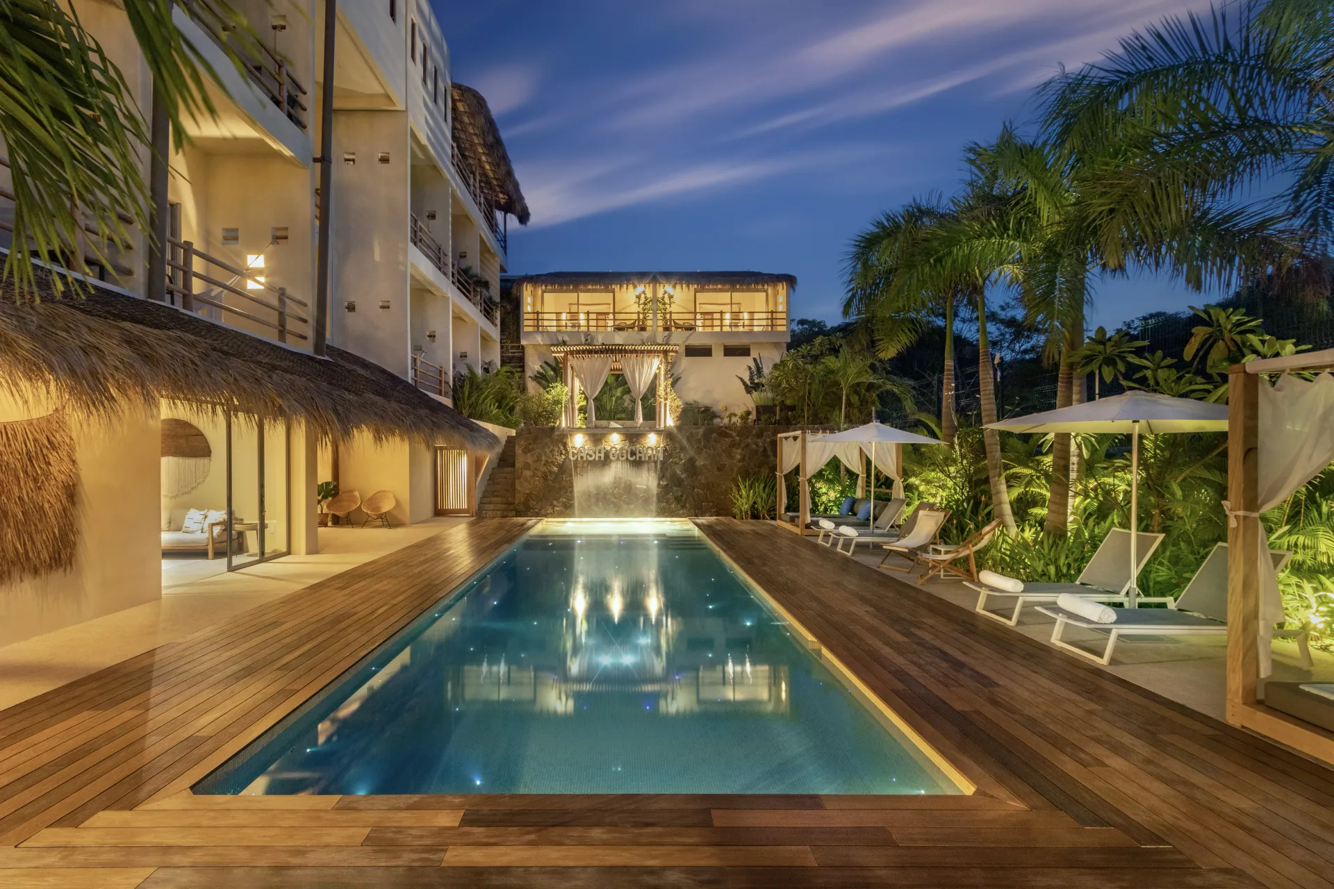 Piscina al atardecer, flanqueada por una terraza de madera, tumbonas y edificios tropicales con palmeras.