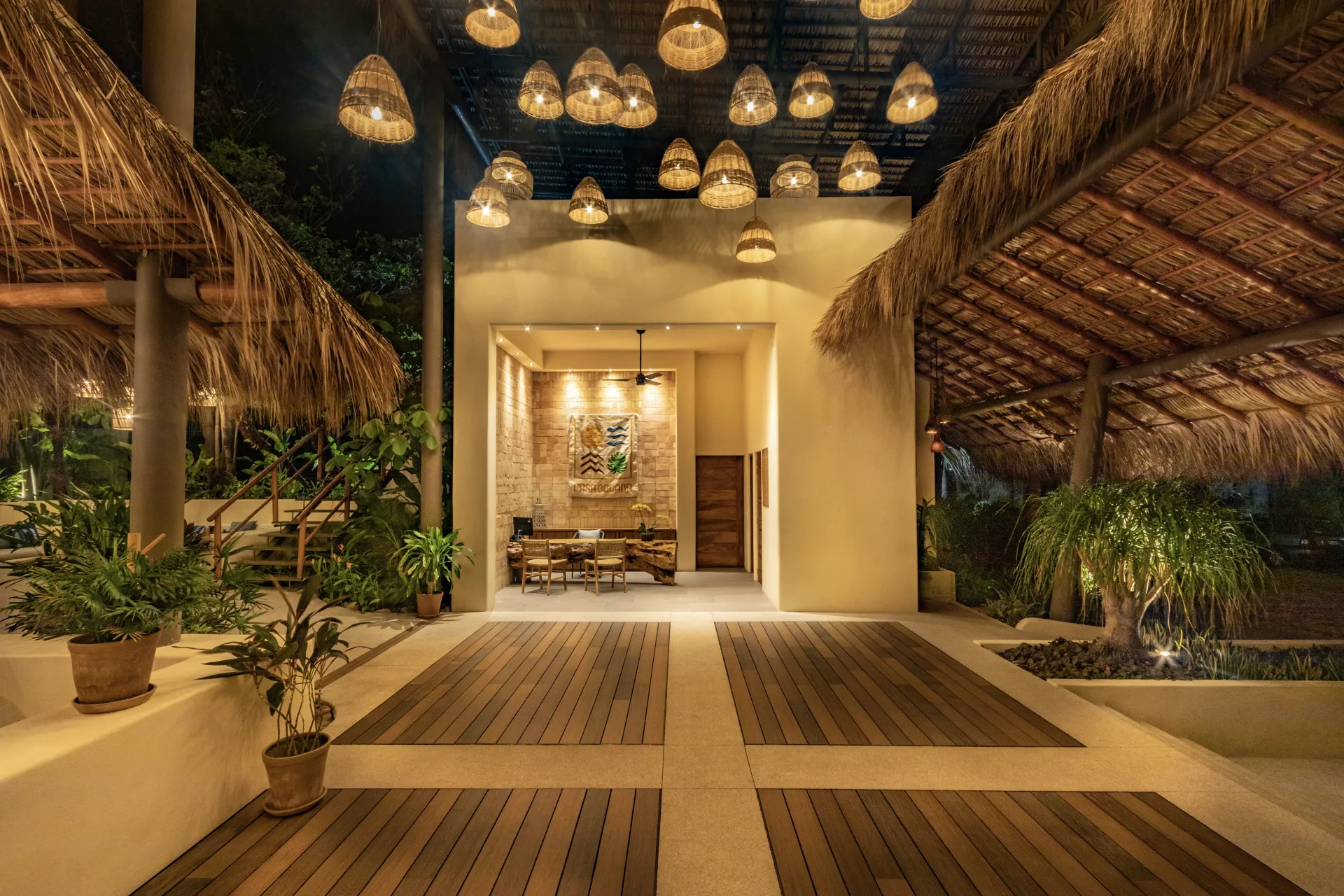 Entrada a un restaurante con techo de paja y lámparas colgantes. Pasarelas de madera y plantas.
