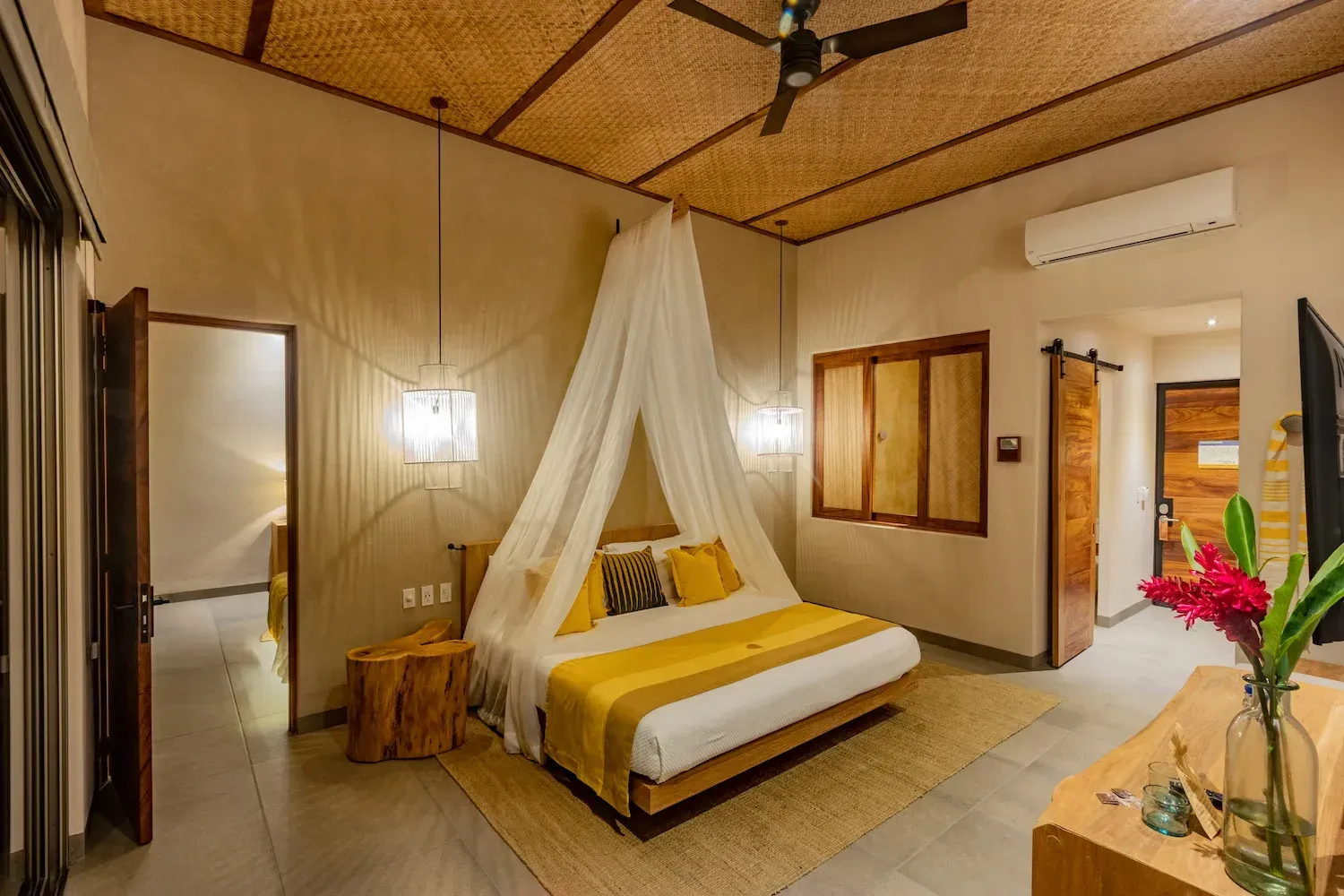 Dormitorio con cama con dosel, detalles amarillos, techo de bambú, puerta abierta al pasillo.