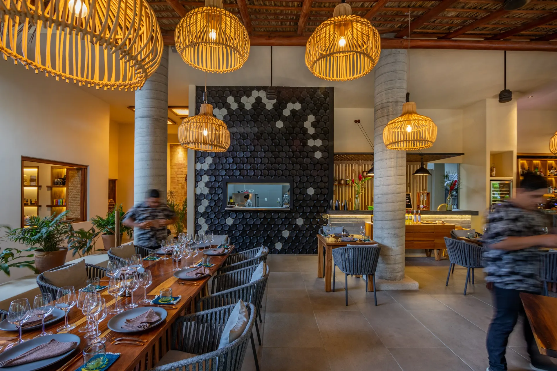 Interior del restaurante: mesa larga preparada para comer, lámparas colgantes de mimbre, y personas desenfocadas.
