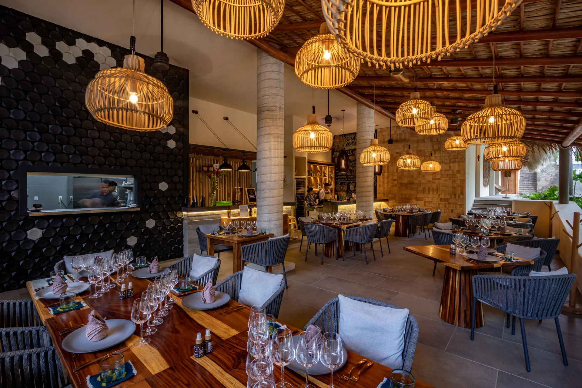 Interior el restaurante con lámparas colgantes de mimbre, mesas dispuestas para comer y cocina abierta.