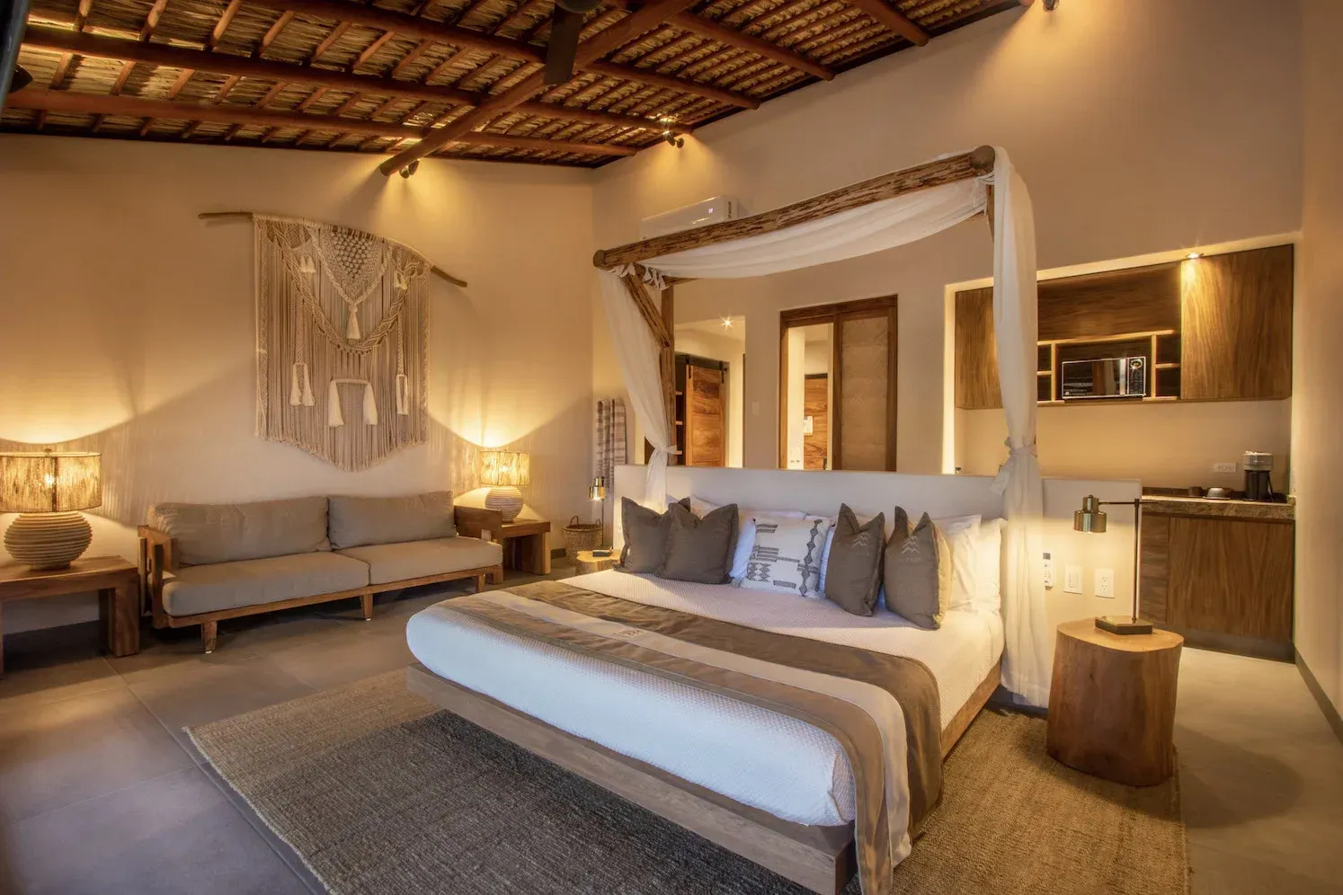 Dormitorio con cama con dosel, sofá y tapiz de pared tejido; tonos neutros, iluminación cálida.