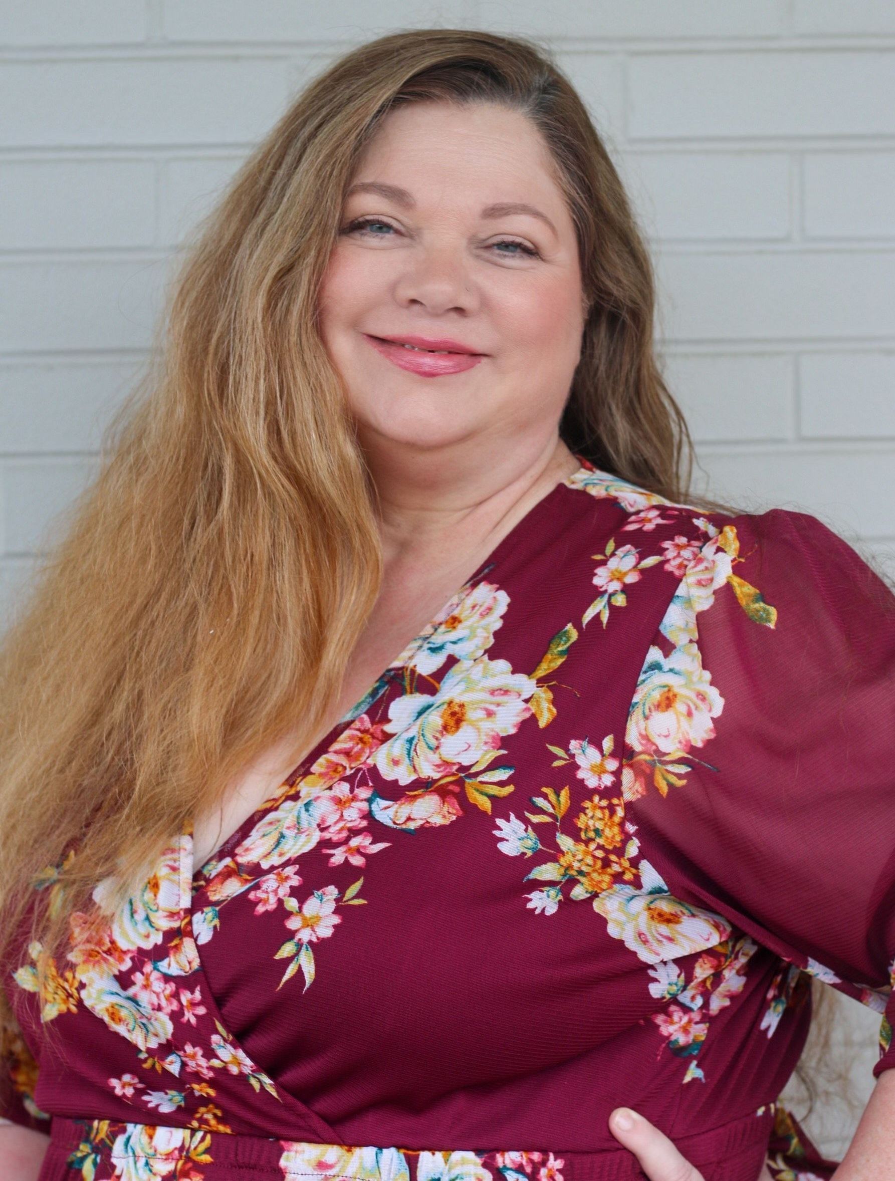 A woman in a maroon floral dress is smiling and standing in front of a white brick wall.