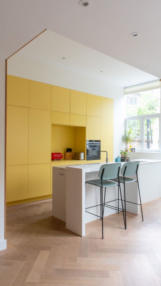 Moderne keuken met gele kastjes, een wit kookeiland met twee turquoise barkrukken en een houten vloer.