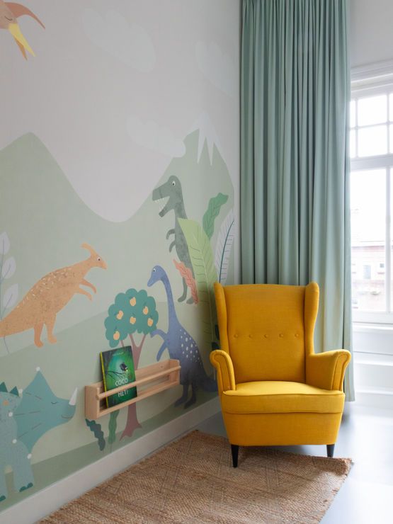 Dinosaurussenmuurschildering in een kinderkamer met een gele fauteuil, boeken, een vloerkleed en groene gordijnen.