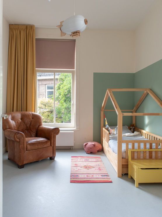 Kinderkamer: bruine fauteuil, huisvormig bed, groene accentmuur, lichtgrijze vloer, raam met uitzicht.