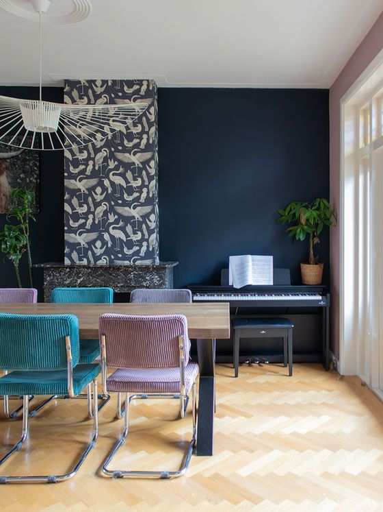 Eetkamer met blauwe muur, piano, kleurrijke stoelen en houten vloer.