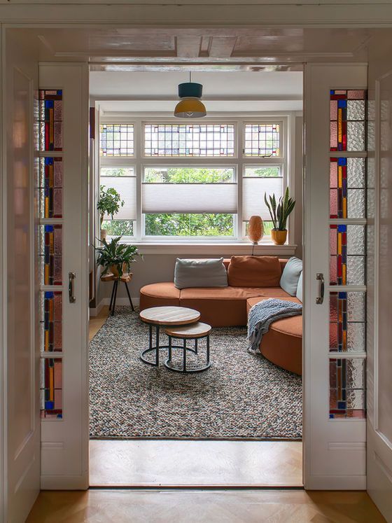 Uitzicht vanuit de woonkamer door de deuropening, met een oranje bank, een vloerkleed met patroon, glas-in-loodramen en planten bij een raam.