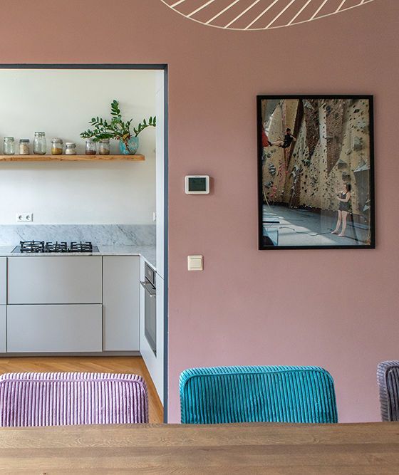 Eetkamer met roze muren en open keuken. Inclusief schilderij, kleurrijke stoelen en een vloerkleed.