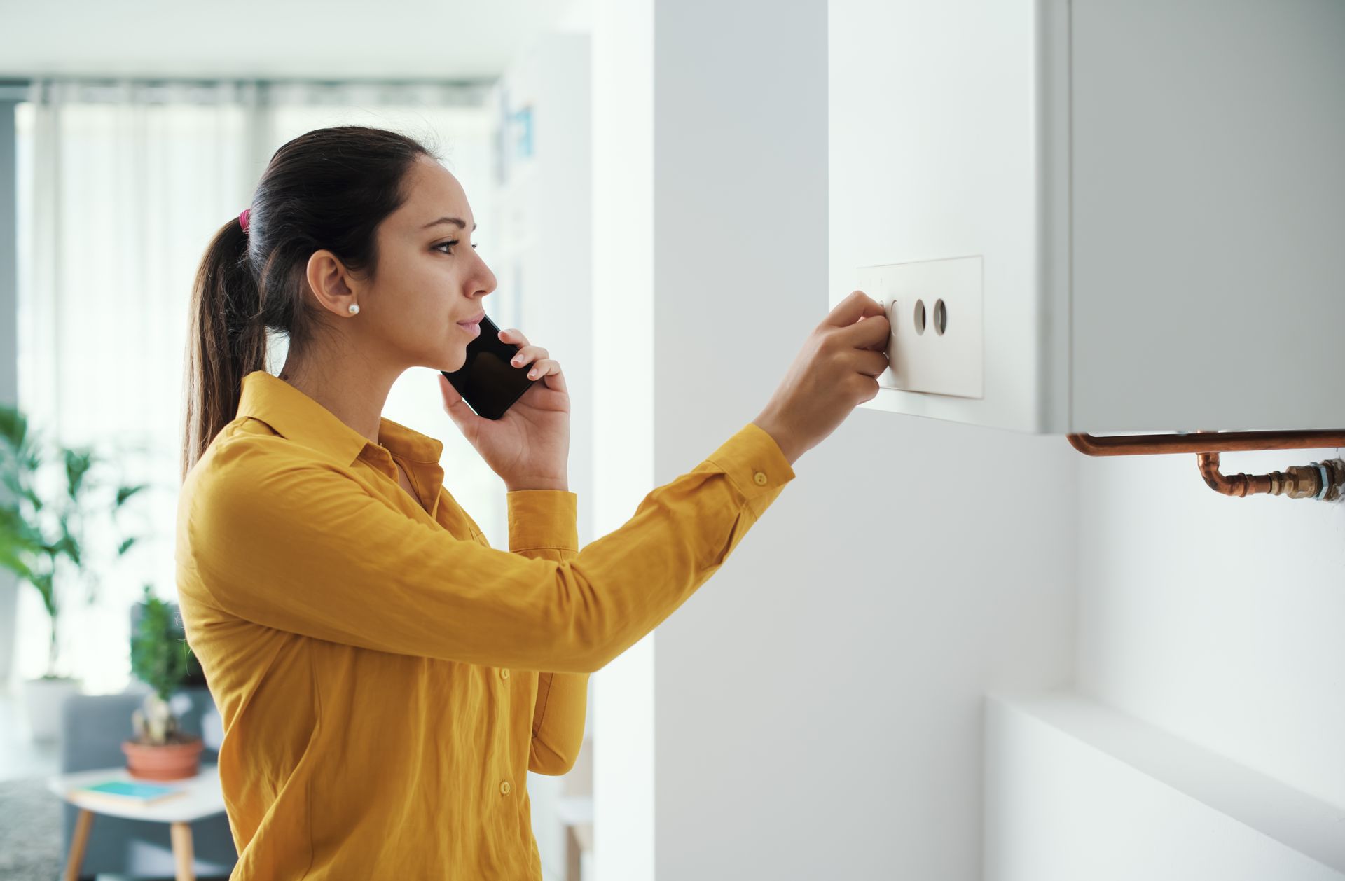 A woman is talking on a cell phone while adjusting a thermostat.