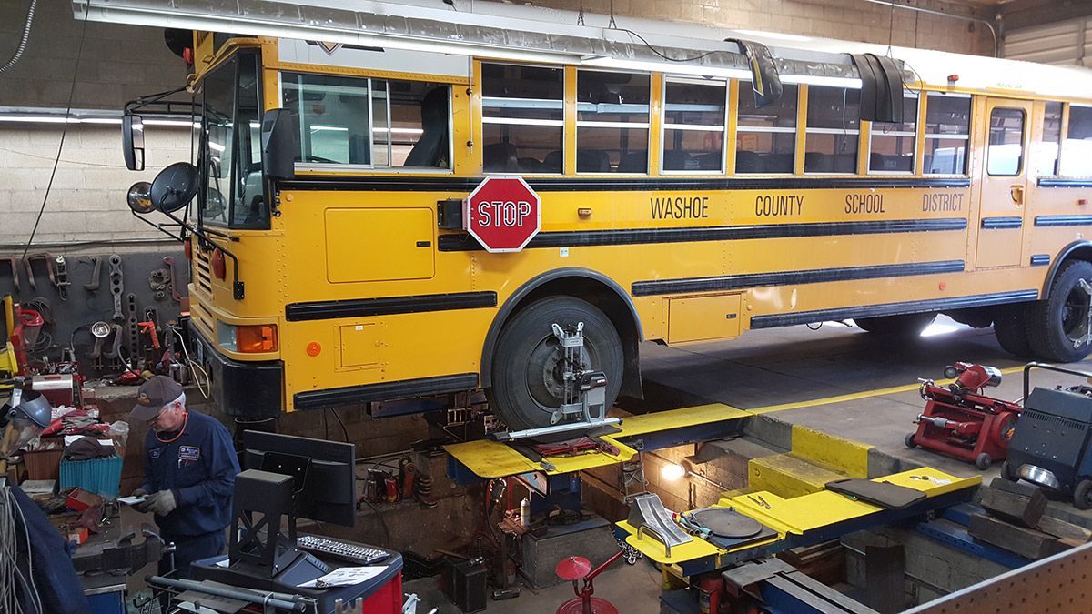 Yellow school bus on a lift in a mechanic’s garage, with a worker below and tools around it | Sunshine Service Automotive Specialists
