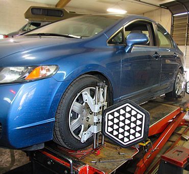 Blue sedan on a wheel alignment rack in a garage, with a calibration target near the front wheel | Sunshine Service Automotive Specialists