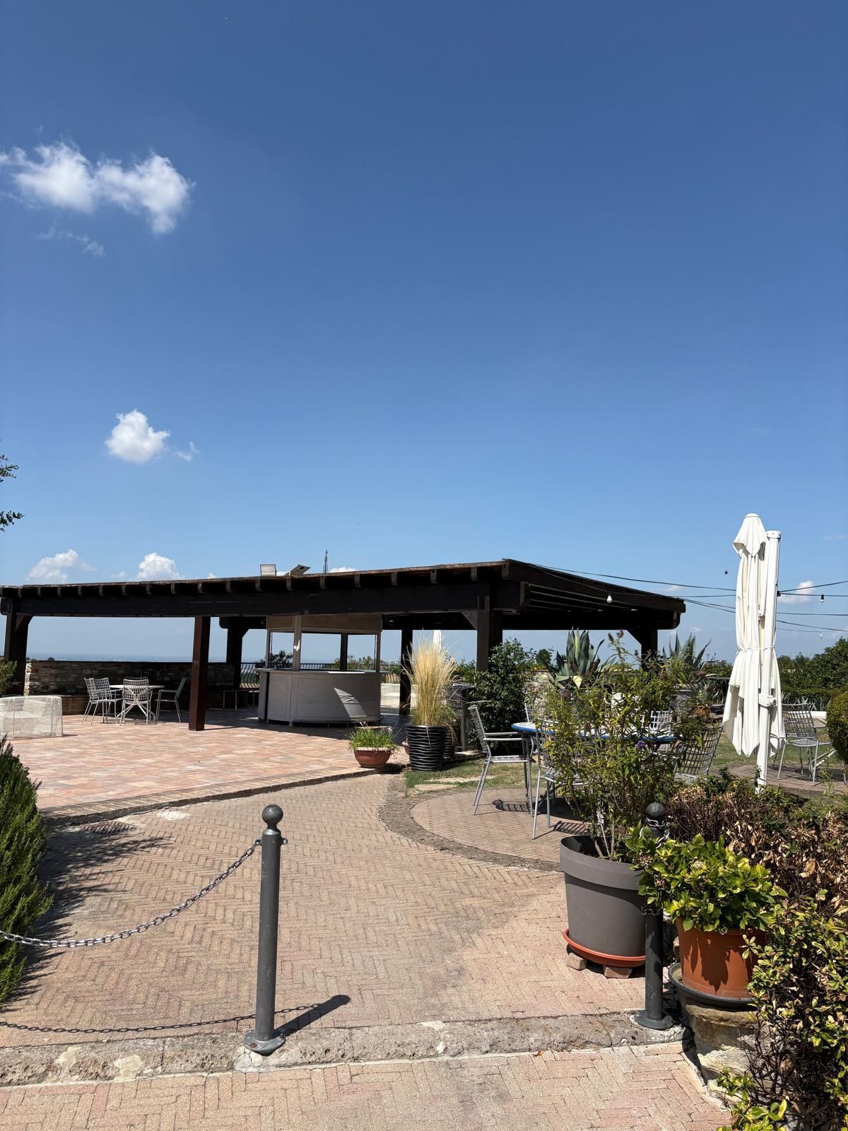 Passerella sul patio con piante in vaso e pergolato ombreggiato sotto un cielo azzurro e limpido.