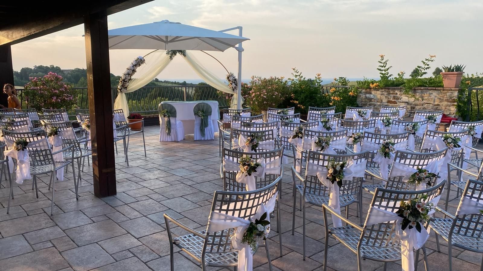 Allestimento per una cerimonia di matrimonio all'aperto con sedie bianche, arco floreale e ombrelloni su un patio in pietra al tramonto.