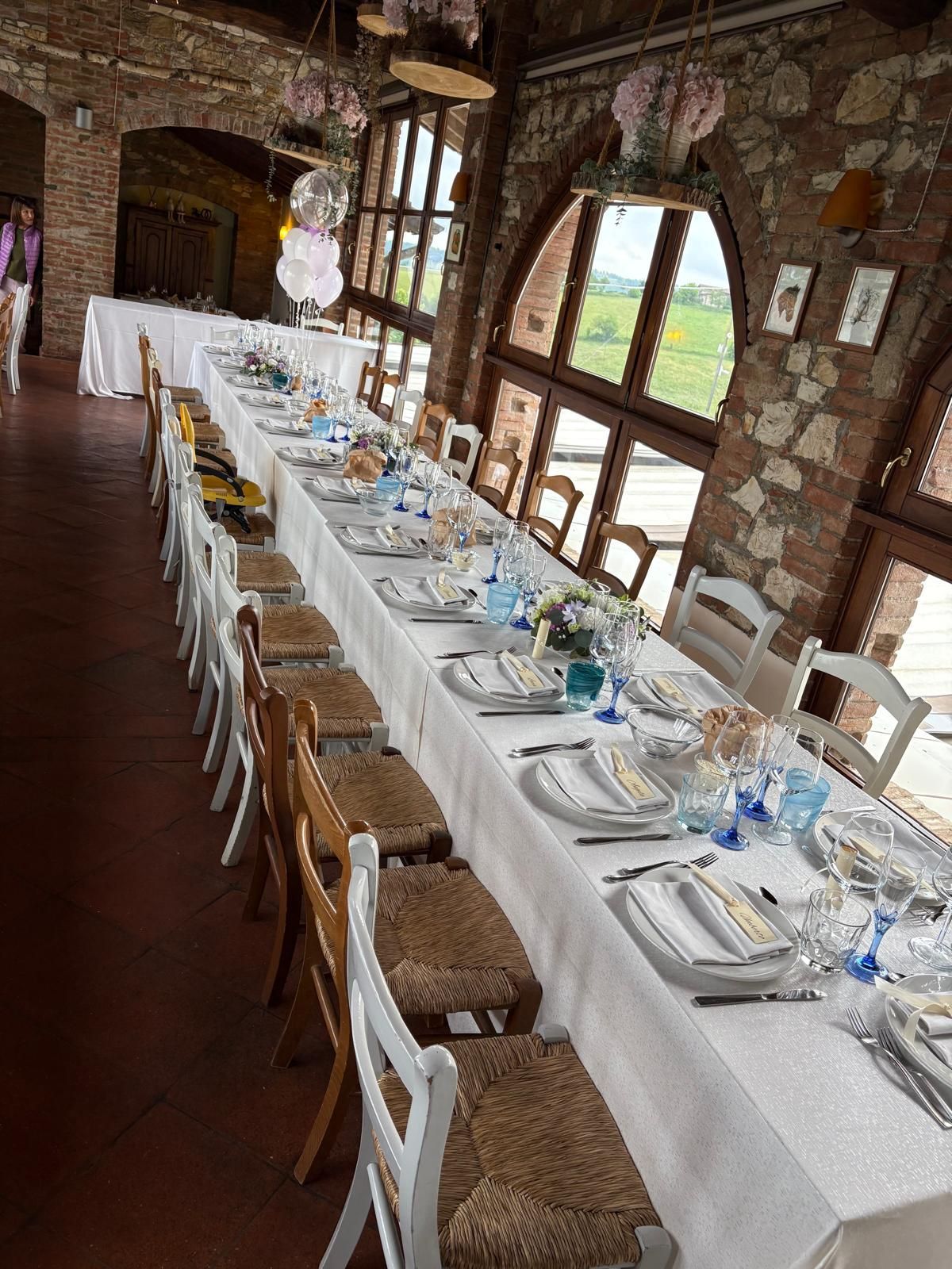 Un lungo tavolo da pranzo apparecchiato con tovaglie e piatti bianchi in una rustica stanza in pietra vicino alle finestre.