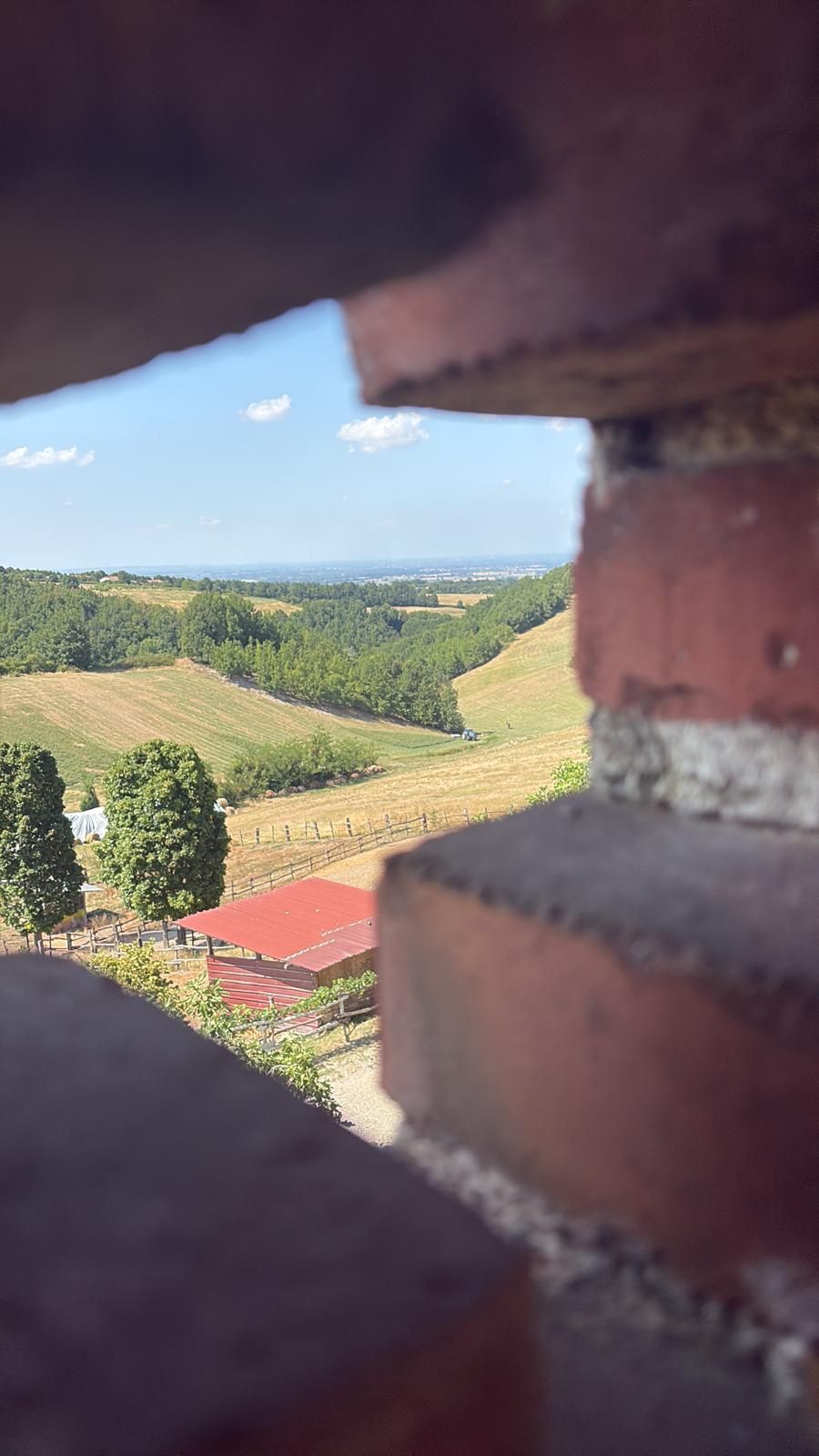 Vista attraverso un'apertura in mattoni di una campagna soleggiata con campi, alberi e una casa dal tetto rosso sotto un cielo azzurro.