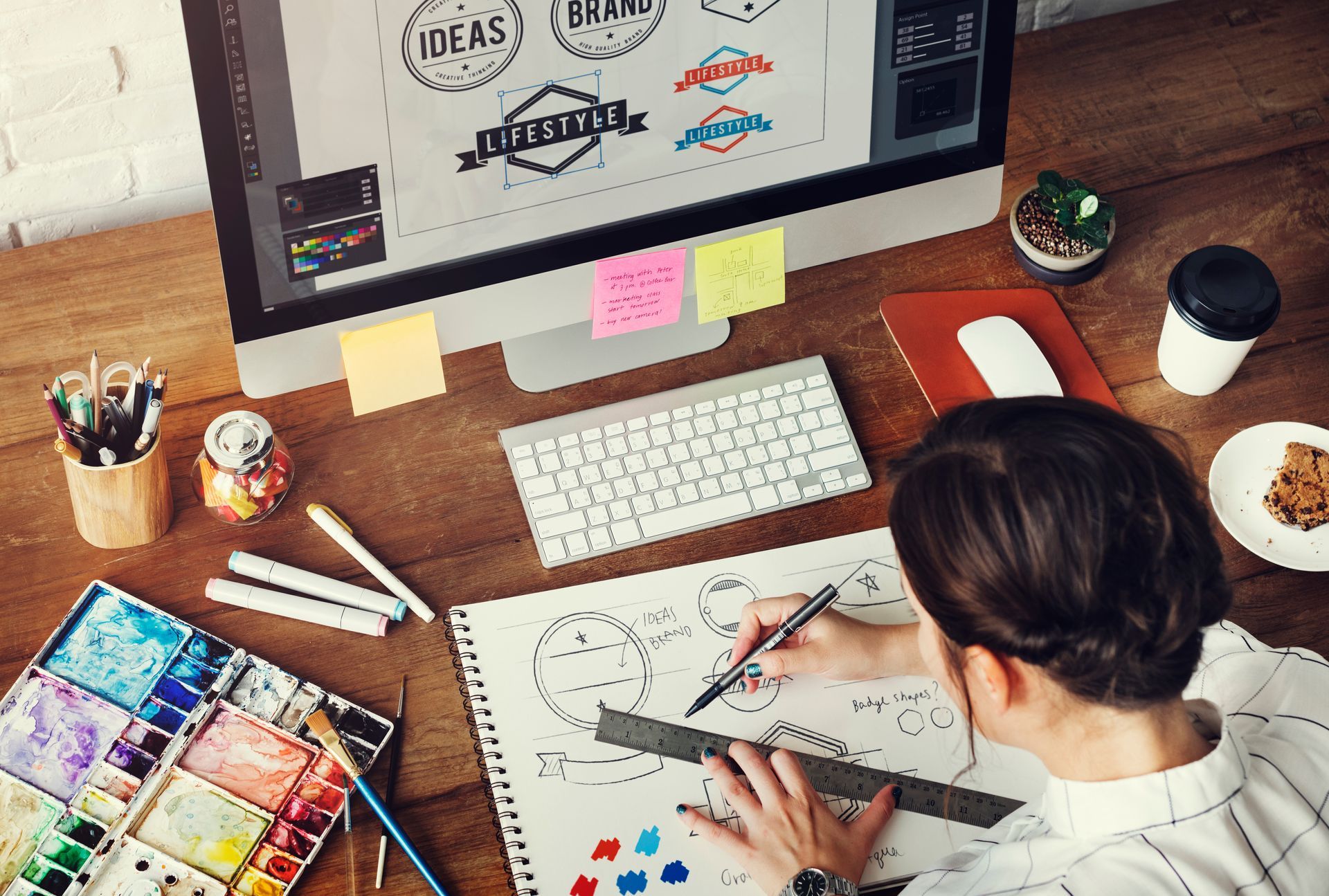 Designer sketching at a desk with laptop, keyboard, color swatches, and sticky notes in a creative workspace