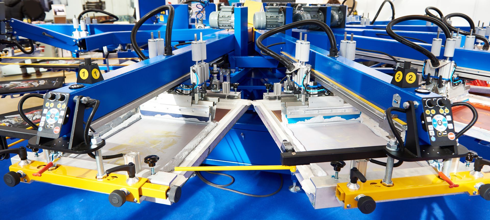 Blue industrial screen-printing press with two white platens and multiple mechanical arms on a factory floor