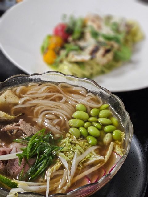 Bowl of noodle soup with edamame and herbs, with a blurred salad plate in the background