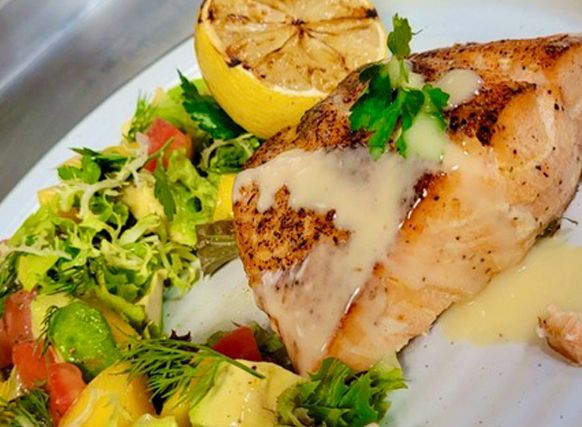 Grilled fish fillet with lemon and mixed greens on a white plate, drizzled with sauce