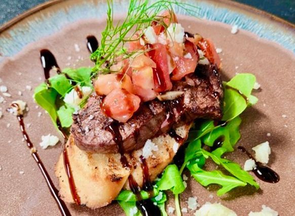 Grilled steak on mashed potatoes with greens, topped with diced tomatoes and herbs on a brown plate