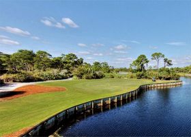 Curved golf green beside dark water, with trees, sand bunker, and blue sky