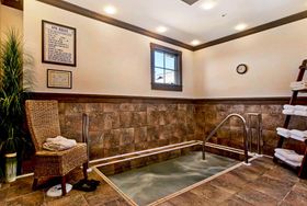 Indoor spa room with tiled soaking pool, metal handrails, wicker chair, and warm stone walls