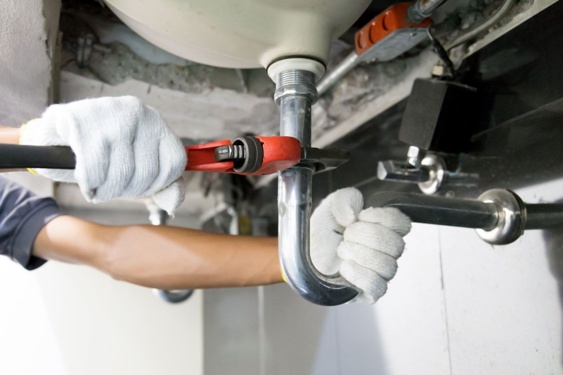 A plumber wearing gloves uses a wrench to tighten a pipe under a sink.