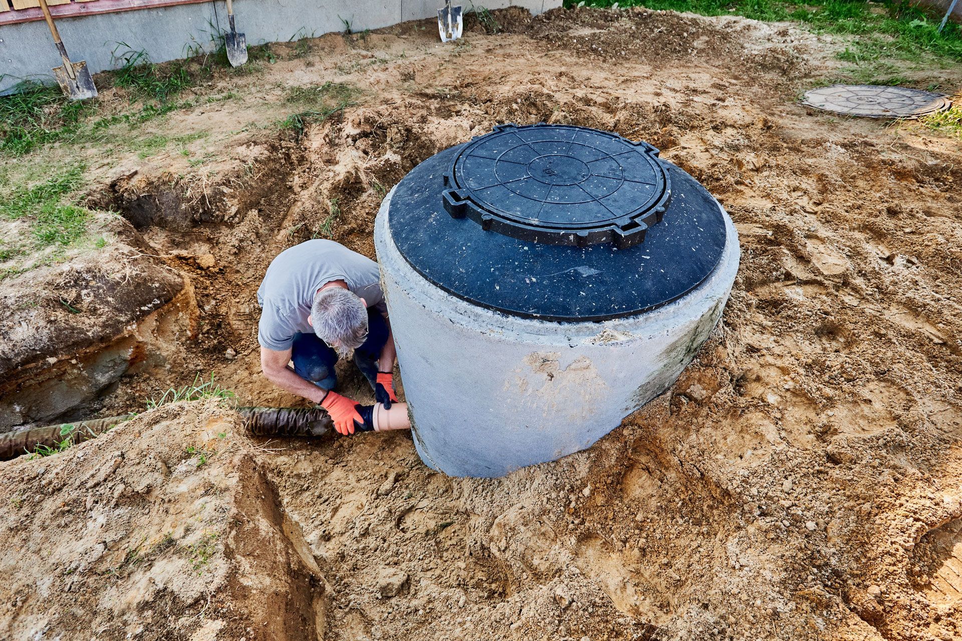 Man installing a pipe connected to a concrete septic tank in a sandy yard. He is wearing gloves.