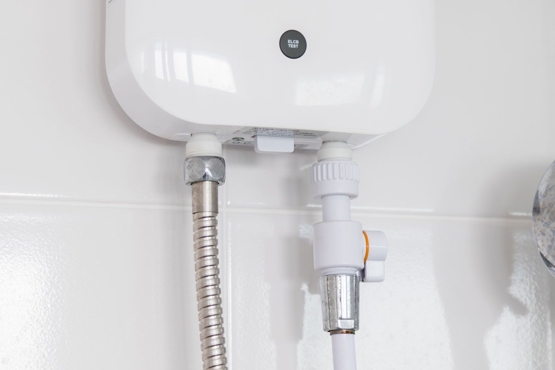White electric water heater mounted on a white tiled wall, with silver and white hoses connected.