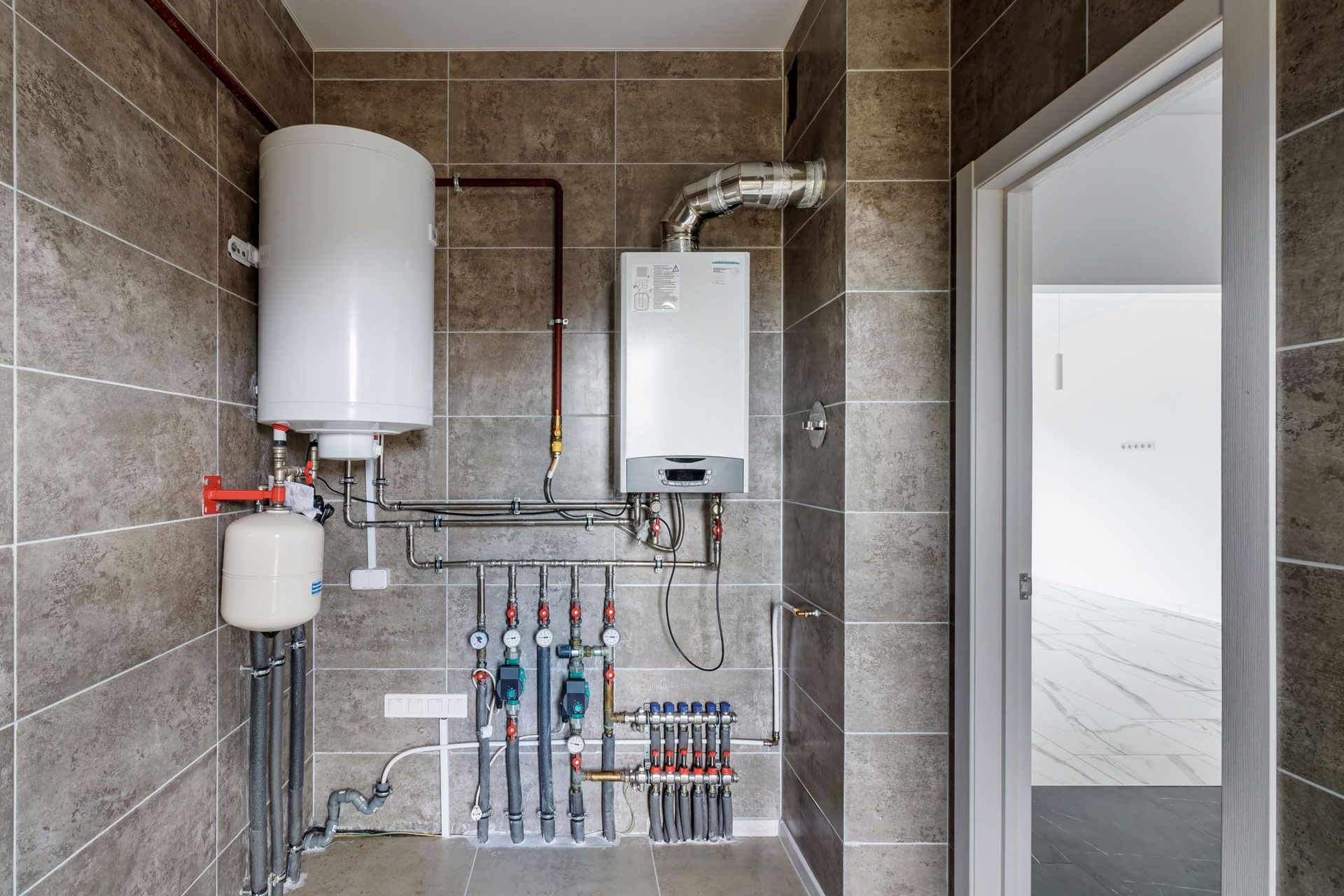 A utility room with a water heater, boiler, and pipes against a gray tiled wall. An open doorway is to the right.