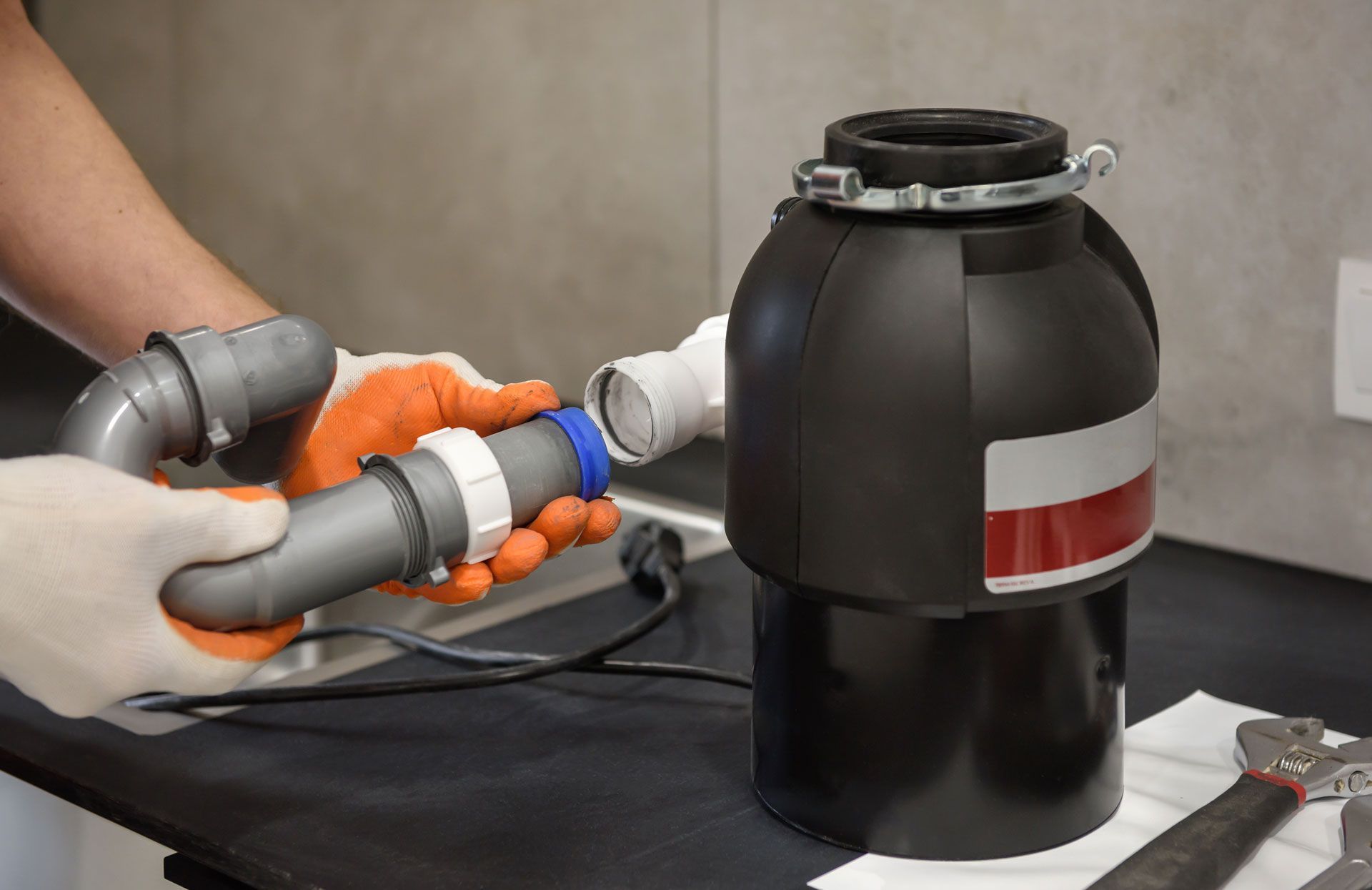 A person with orange gloves connects plumbing to a garbage disposal on a countertop.