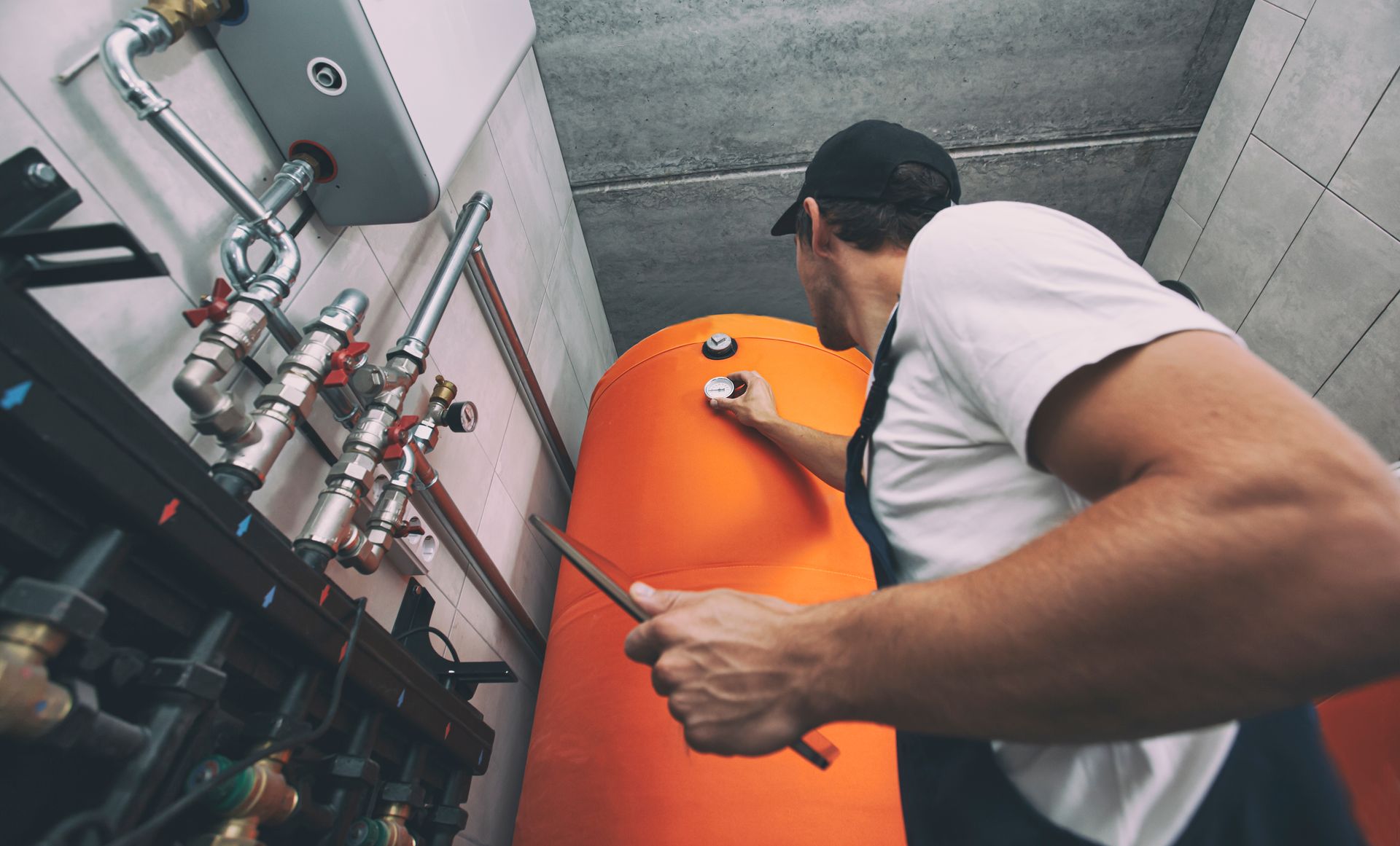 Plumber checks an orange water heater in a utility room, pointing with a screwdriver.